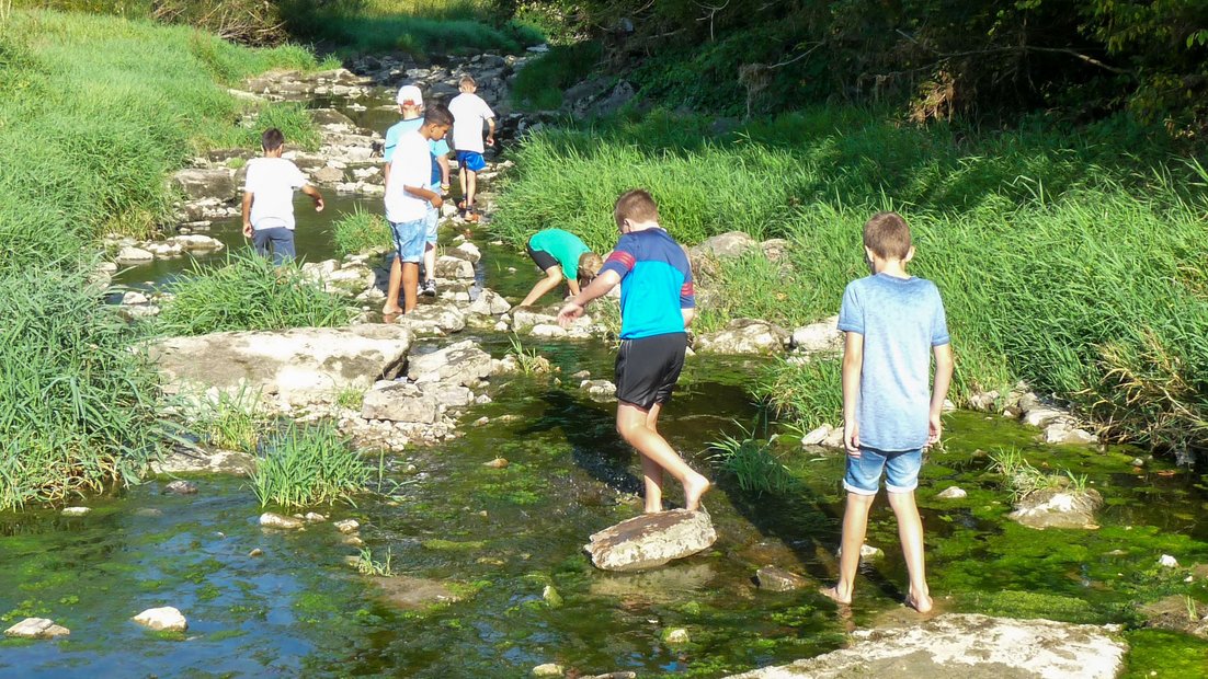 Gli studenti più giovani esplorano un letto di fiume poco profondo con grandi pietre durante una giornata di escursioni.
