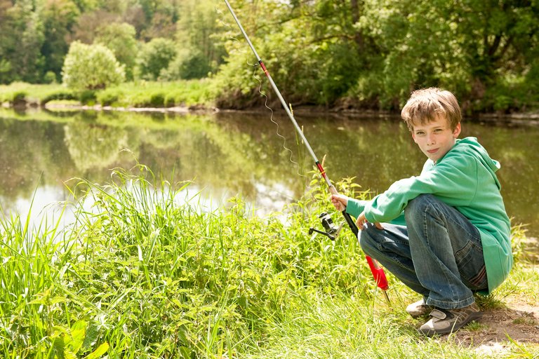Il ragazzo si siede sulla riva del fiume con una canna da pesca in mano.